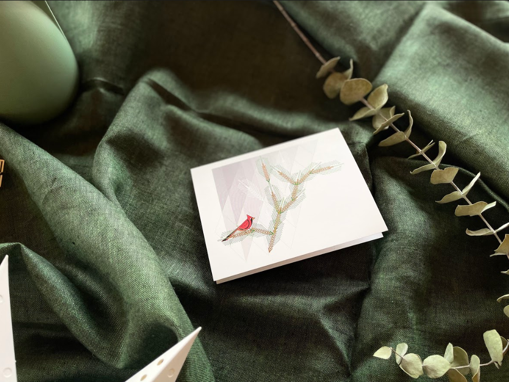 Holiday greeting card depicting an abstracted cardinal design. Pictured on a green fabric background. Recommended in the 2021 Gift Guide.